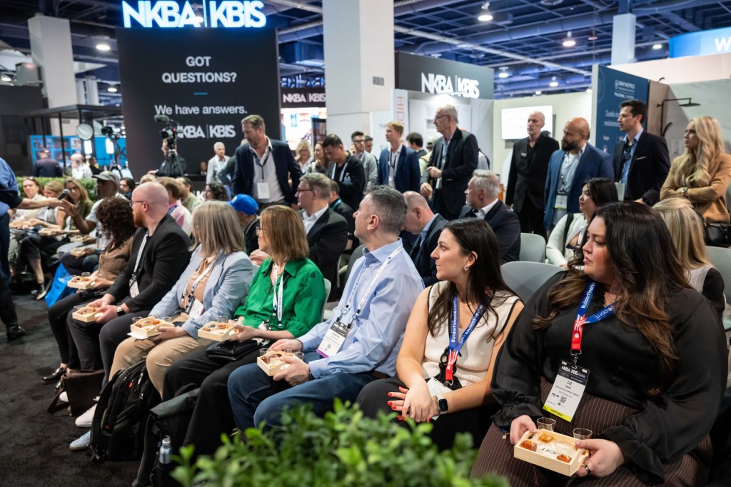 A diverse audience seated and engaged at a conference, with some attendees holding food. A large display in the background features the text 'GOT QUESTIONS? We have answers.'