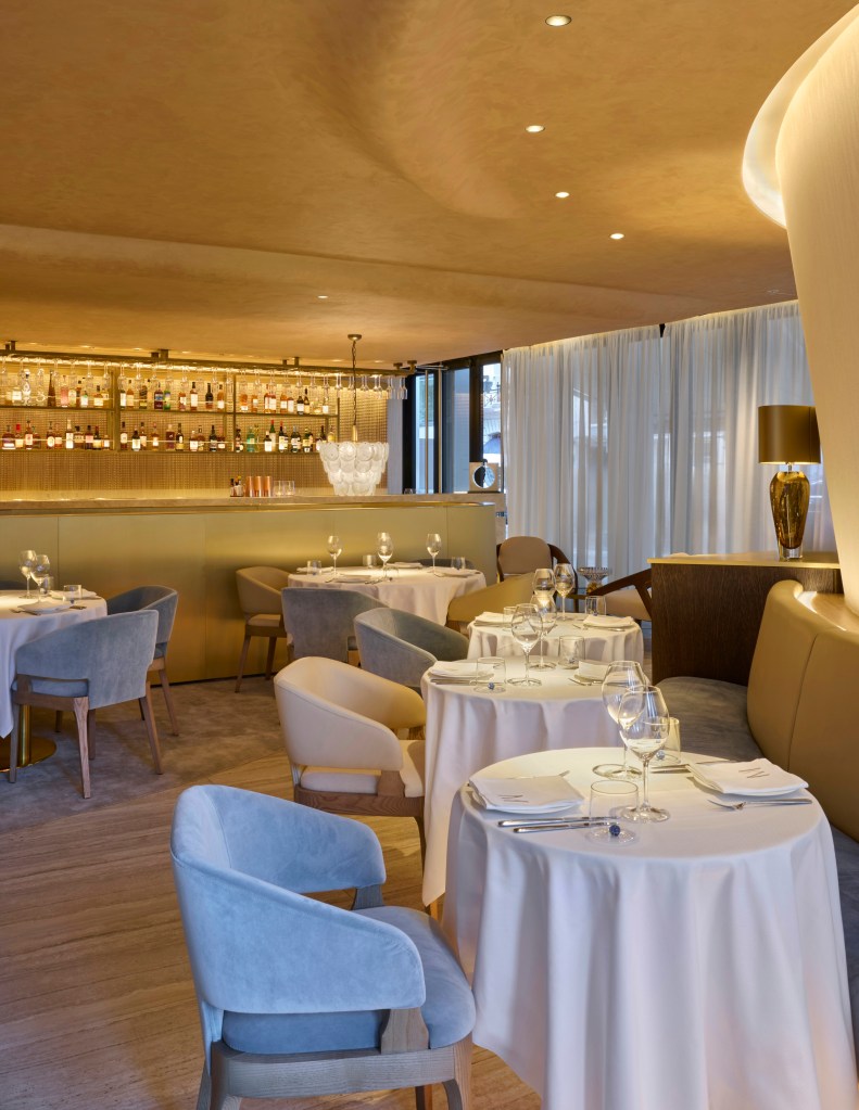 Elegant restaurant interior featuring neatly set tables with white tablecloths, stylish chairs in soft hues, and a well-stocked bar in the background with ambient lighting.