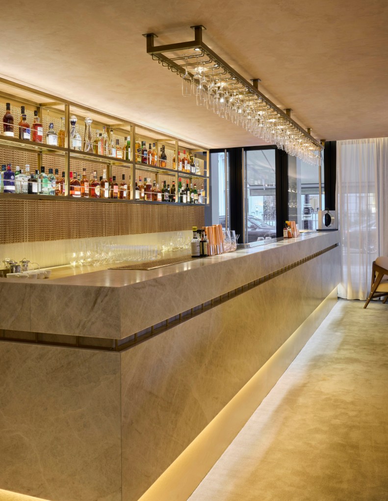 A modern bar with a sleek marble countertop, illuminated by warm lighting and lined with various alcoholic beverages on shelves above.