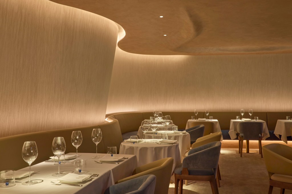 Interior of a modern restaurant featuring curved walls, soft lighting, elegant dining tables with tableware, and comfortable chairs.