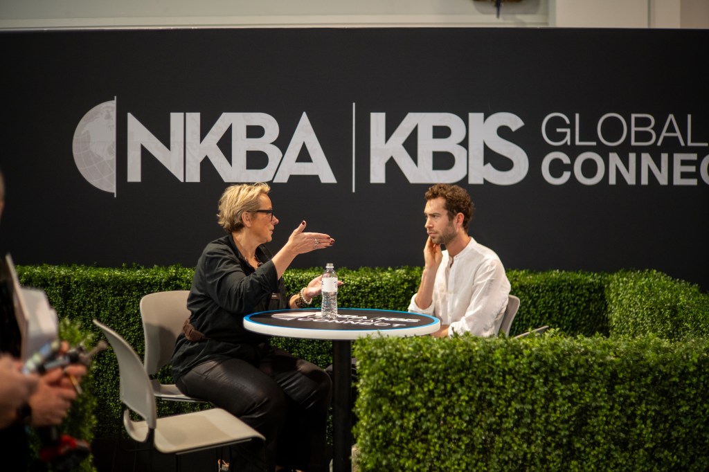 A woman and a man engage in conversation at a round table, surrounded by greenery, with a backdrop featuring the NKBA and KBIS Global Connect logos.