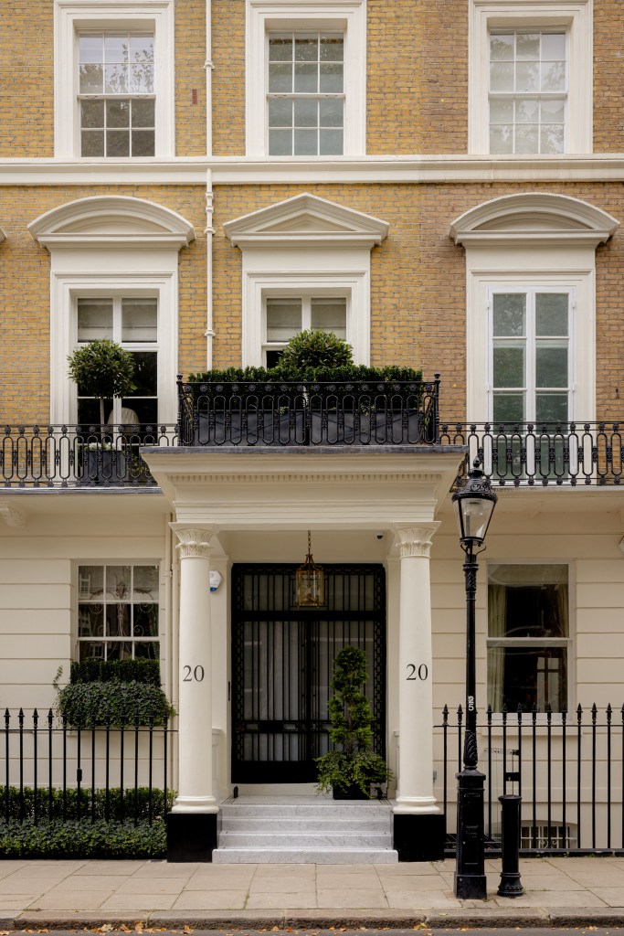 Front view of an elegant townhouse with a black wrought-iron railing, large windows, and a decorative entrance featuring marble steps and two potted plants flanking the door.