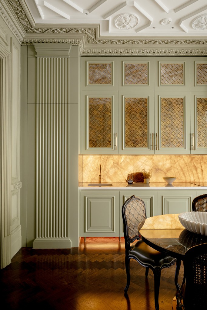 Elegant kitchen interior featuring green cabinetry with glass detailing, decorative ceiling, and a stylish dining table with black chairs.