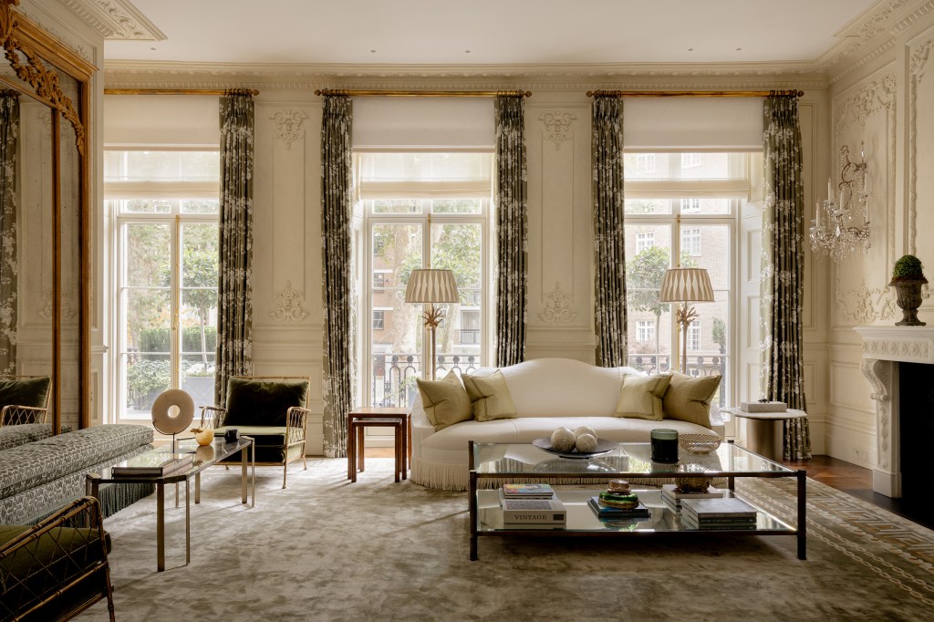 A stylish living room featuring elegant décor, large windows with flowing curtains, a comfortable sofa with decorative cushions, a glass coffee table with various items, and a rich carpet, creating a sophisticated atmosphere.