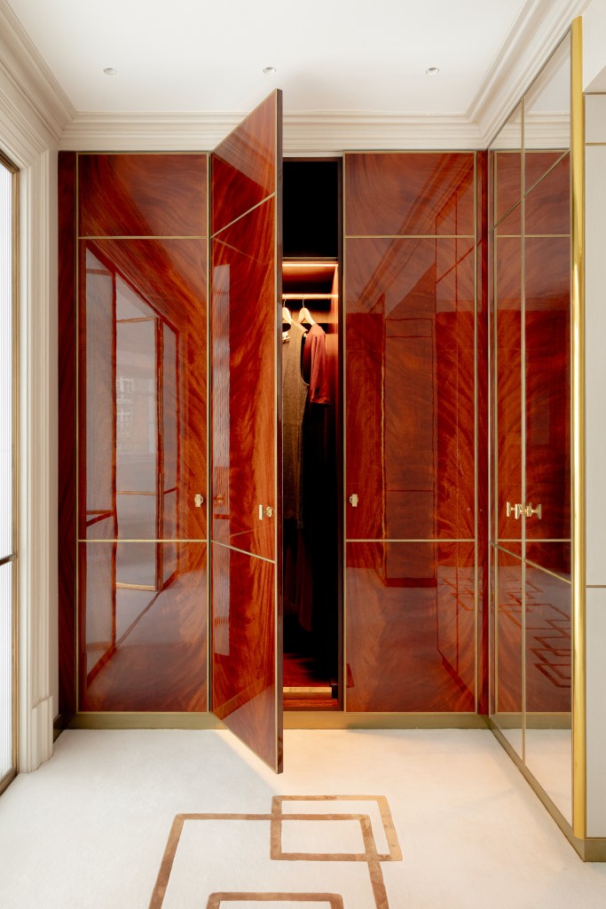 A luxurious walk-in wardrobe with polished wooden doors, partially open to reveal hanging clothes, featuring elegant brass accents and soft lighting.