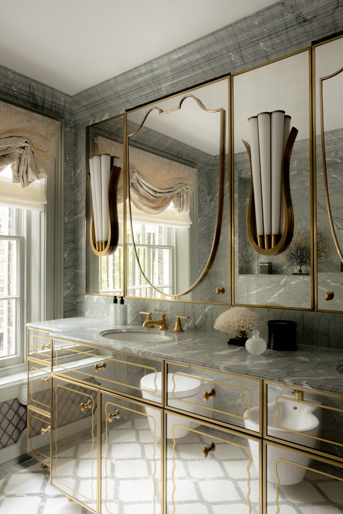 A stylish bathroom featuring grey marble walls, a reflective vanity with gold detailing, and contemporary lighting fixtures. It includes a round mirror above the sink, decorative elements, and large windows allowing natural light.