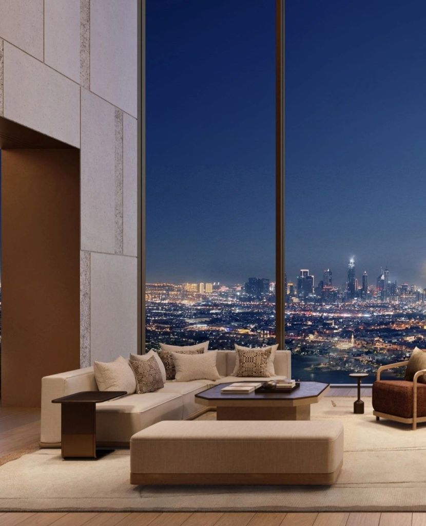 A modern living room featuring a stylish beige sofa with decorative cushions, a coffee table, and a cozy armchair, set against a large window showcasing a panoramic night view of a city skyline.