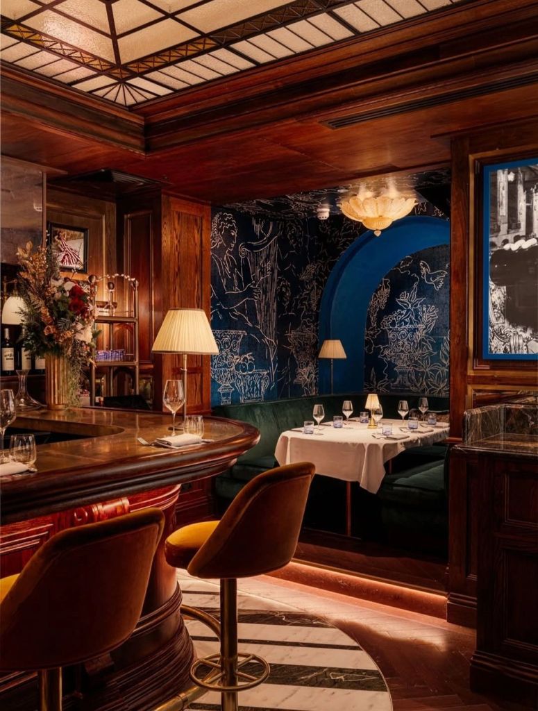 A stylish interior of a restaurant featuring a wooden bar, velvet seating, and artistic wallpaper. The space includes soft lighting from lamps and a decorative ceiling with stained glass elements.