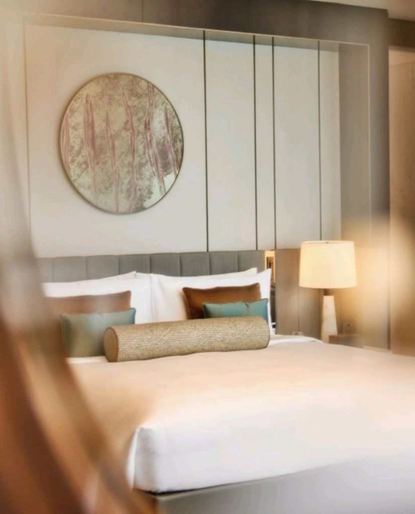 A modern hotel room featuring a neatly made bed with decorative cushions, a round mirror on the wall, and a table lamp beside the bed.