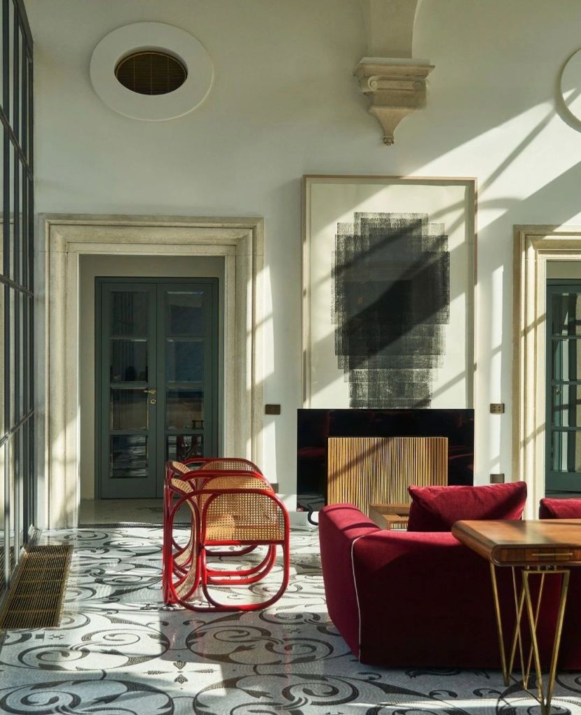 A stylish living room featuring a red sofa, wooden furniture, and a combination of patterned tiles. Large windows allow natural light to illuminate the space, highlighting a piece of abstract artwork on the wall.