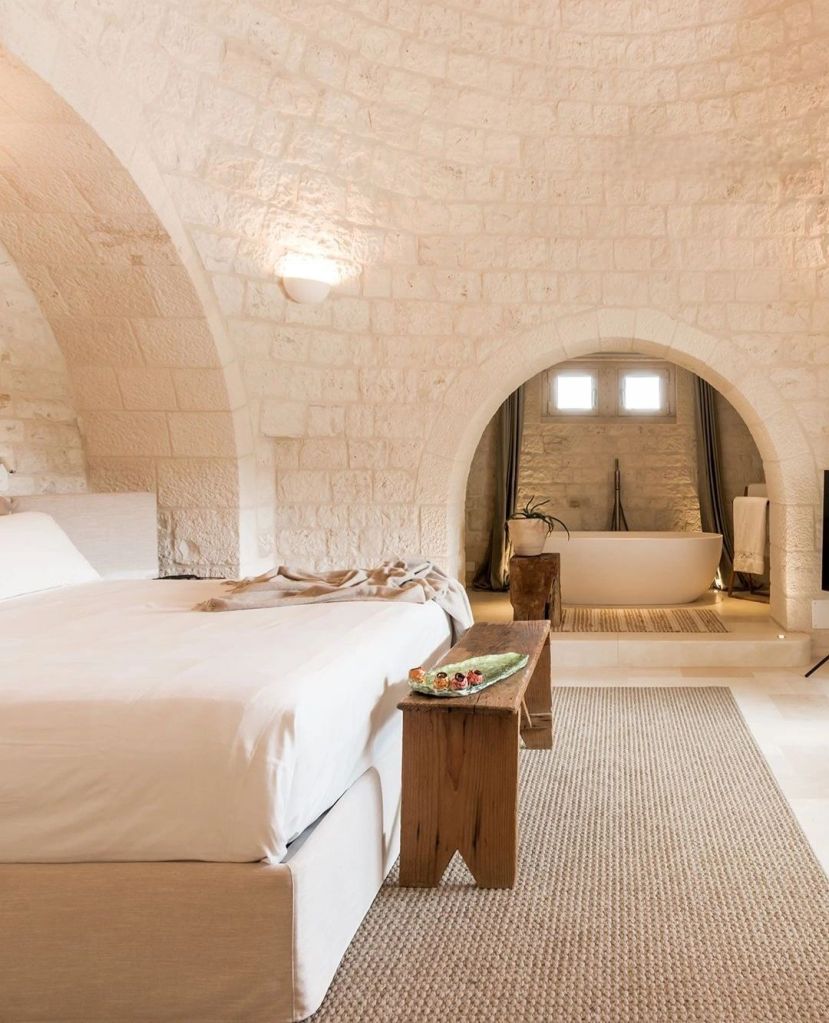 A stylish, minimalist bedroom featuring a large bed with white linens, an arched stone ceiling, and a wooden bench with decorative items. A modern bathtub is visible in the background, complemented by soft lighting and natural elements.