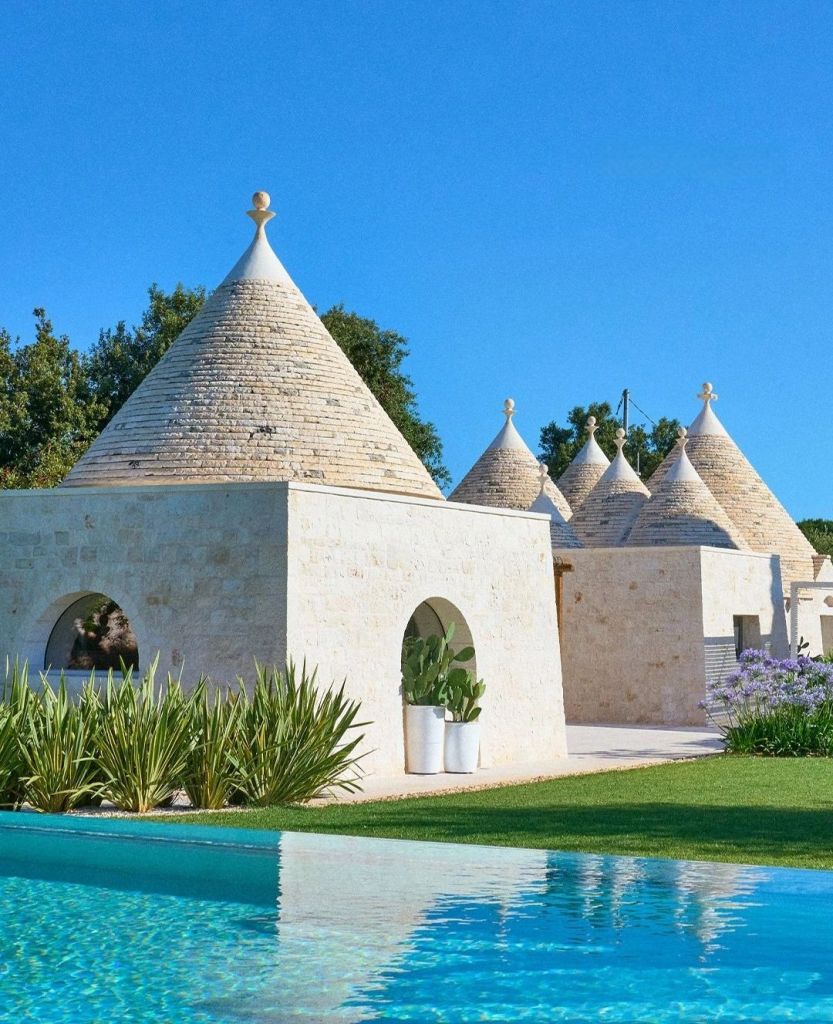 A picturesque view of traditional trullo houses with conical roofs, surrounded by lush greenery and a sparkling swimming pool under a clear blue sky.