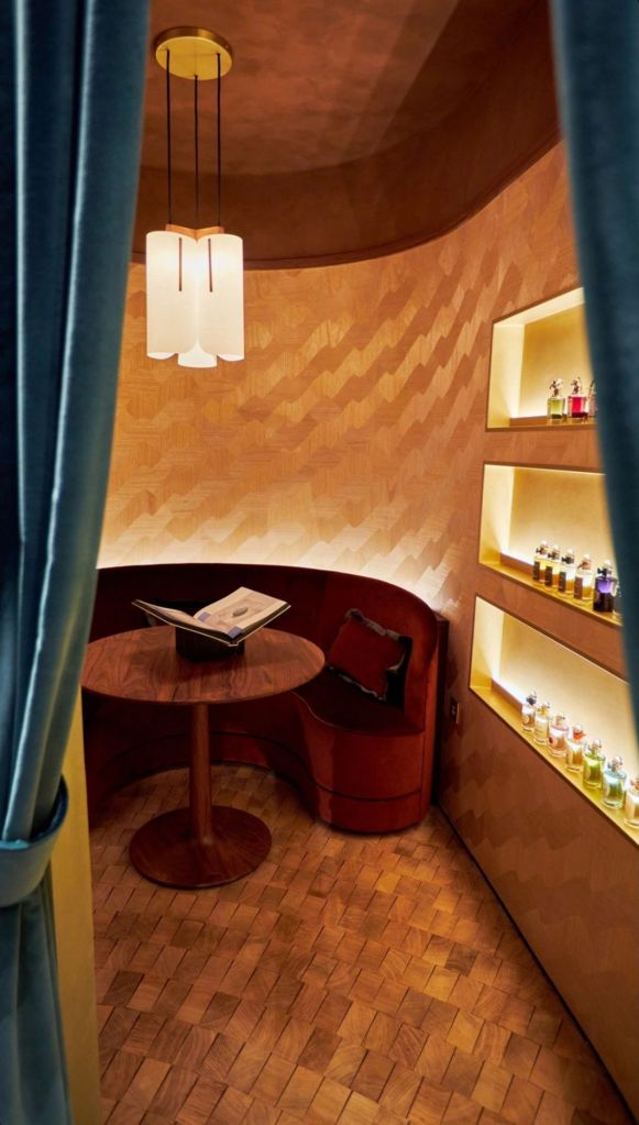 A cosy room featuring a curved wooden seating area, a round table, and a book, with soft lighting and shelves displaying various perfume bottles.