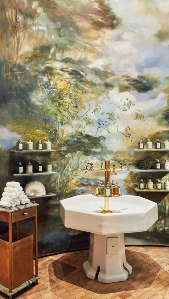 A tranquil spa setting featuring a decorative wall mural of lush greenery and soft clouds, a white octagonal basin in the centre with golden taps, shelves displaying various bottles, and a wooden cart with neatly rolled towels.