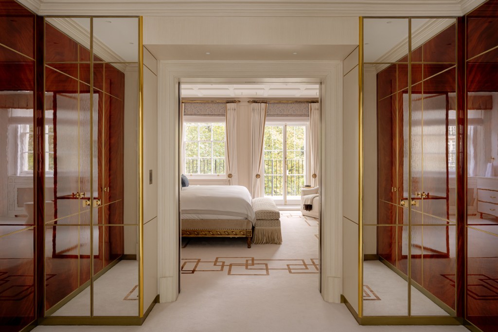 A view of an elegantly designed bedroom through a corridor, featuring mirrored wardrobe doors with gold accents, a large bed with soft bedding, and natural light streaming in from a window adorned with curtains.