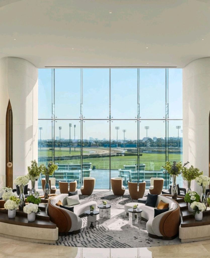 Spacious and modern lounge area with large glass windows overlooking a racetrack, featuring stylish furniture and floral arrangements.