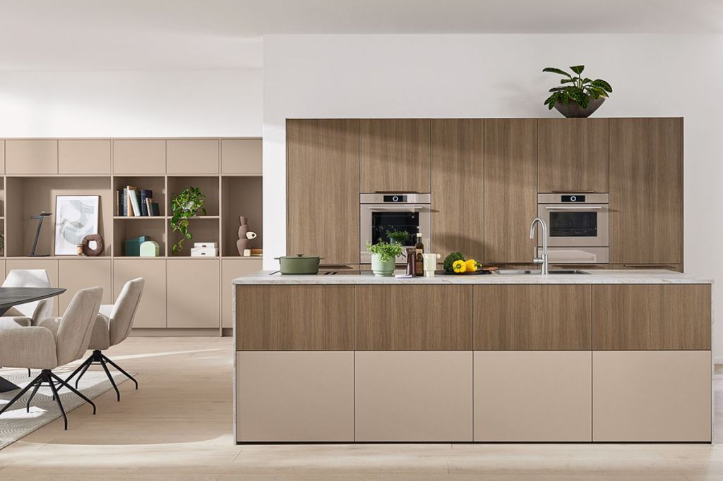 A modern kitchen with a central island featuring a sink and some fruits, surrounded by wooden cabinetry and built-in ovens. The background includes a dining area with a round table and upholstered chairs, and a shelf with decorative items and plants.