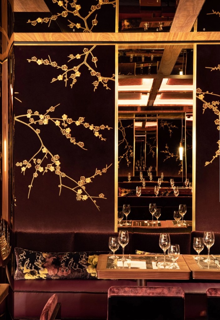 A luxurious restaurant interior featuring dark velvet walls adorned with golden cherry blossom motifs, mirrored panels, and elegantly set dining tables with wine glasses.