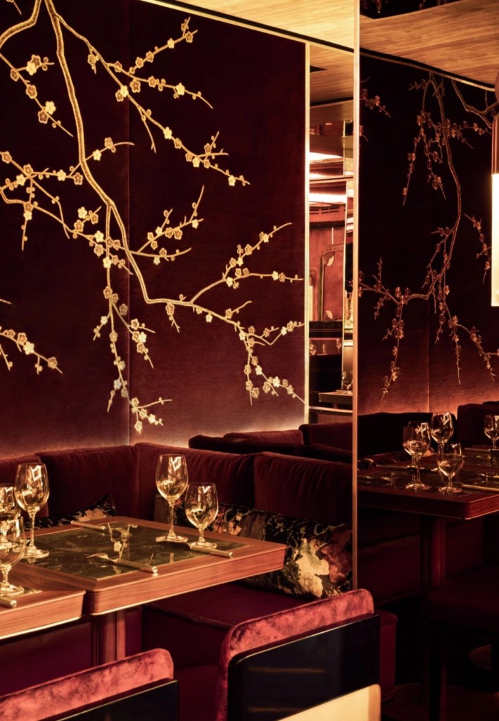 Elegant interior of a restaurant featuring burgundy walls adorned with gold cherry blossom motifs and plush seating, set with glassware on wooden tables.