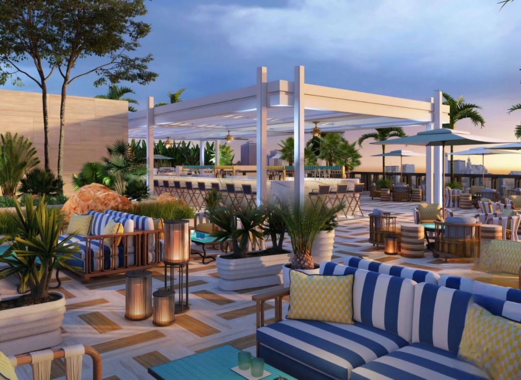 An upscale rooftop lounge with stylish outdoor seating, featuring striped sofas, potted plants, and a bar area under a canopy. The atmosphere is enhanced by warm lighting and a scenic view of the city skyline against a twilight sky.