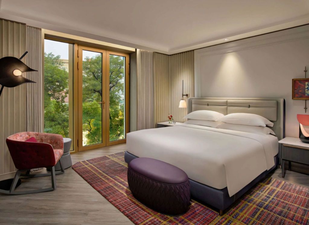 A stylish hotel room featuring a large bed, a patterned rug, a comfortable chair, and large windows with views of greenery.