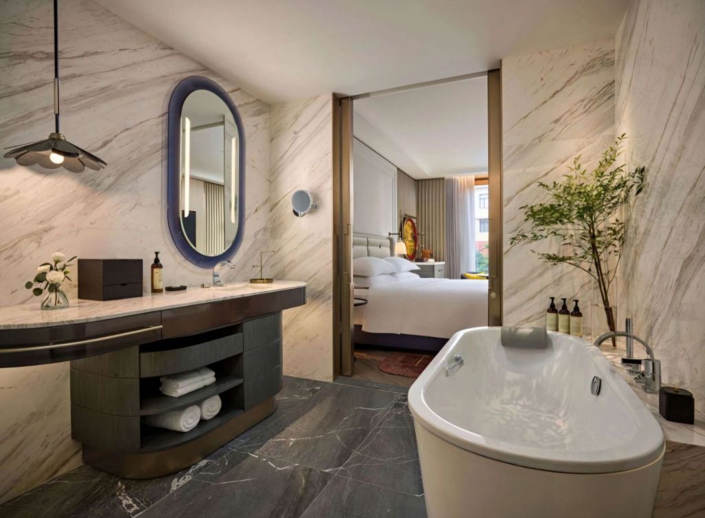 A modern bathroom featuring a freestanding bathtub, a stylish vanity with a large mirror, and marble walls. There is an open doorway leading to a bright bedroom in the background, adorned with curtains and decorative items.