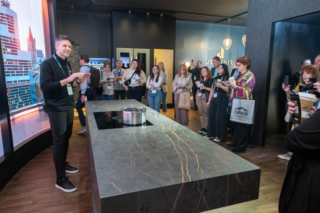 A presenter engages with an audience in a modern kitchen showroom, showcasing a cooking demonstration with a stylish stovetop and a large pot on display.