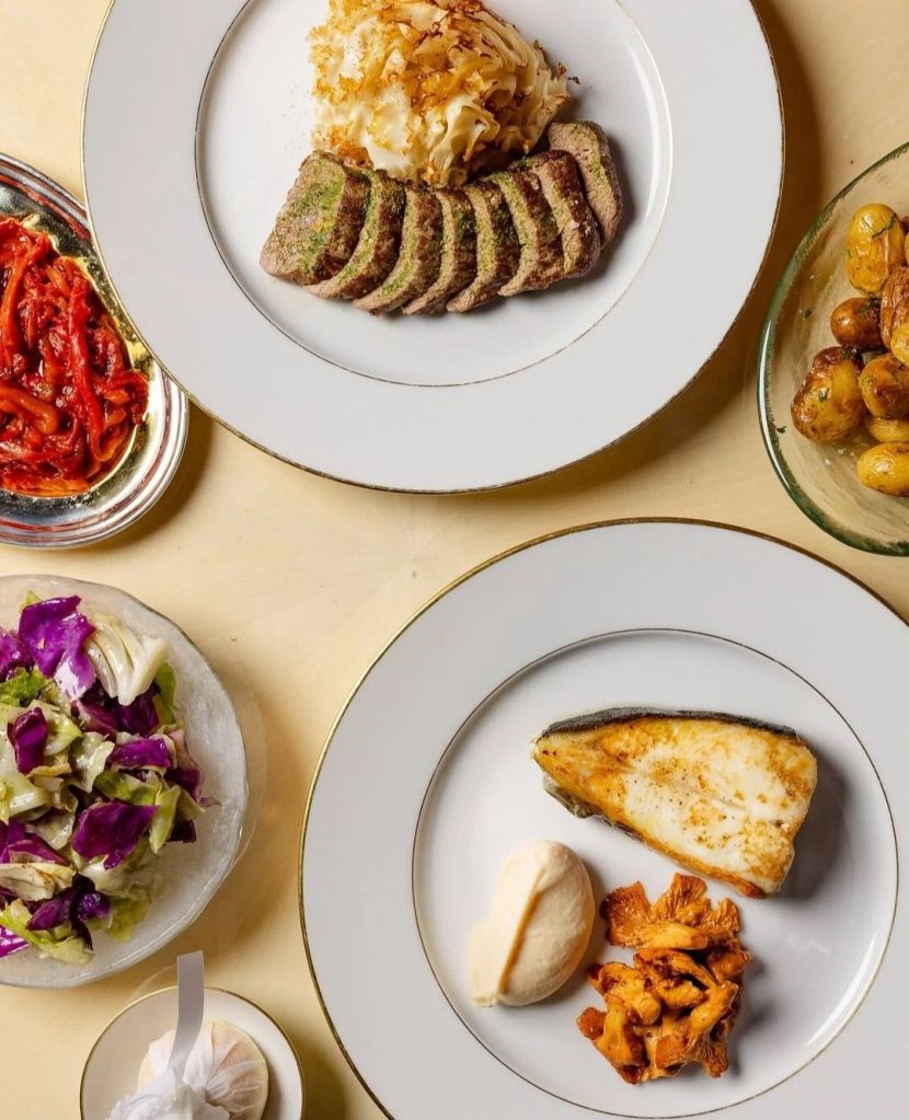 A neatly plated meal featuring slices of meatloaf with herbs, buttery mashed potatoes topped with crispy onions, accompanied by red pepper relish, roasted baby potatoes, and a side salad with purple and green leaves. Another plate includes a piece of pan-seared fish, a dollop of creamy sauce, and sautéed mushrooms.