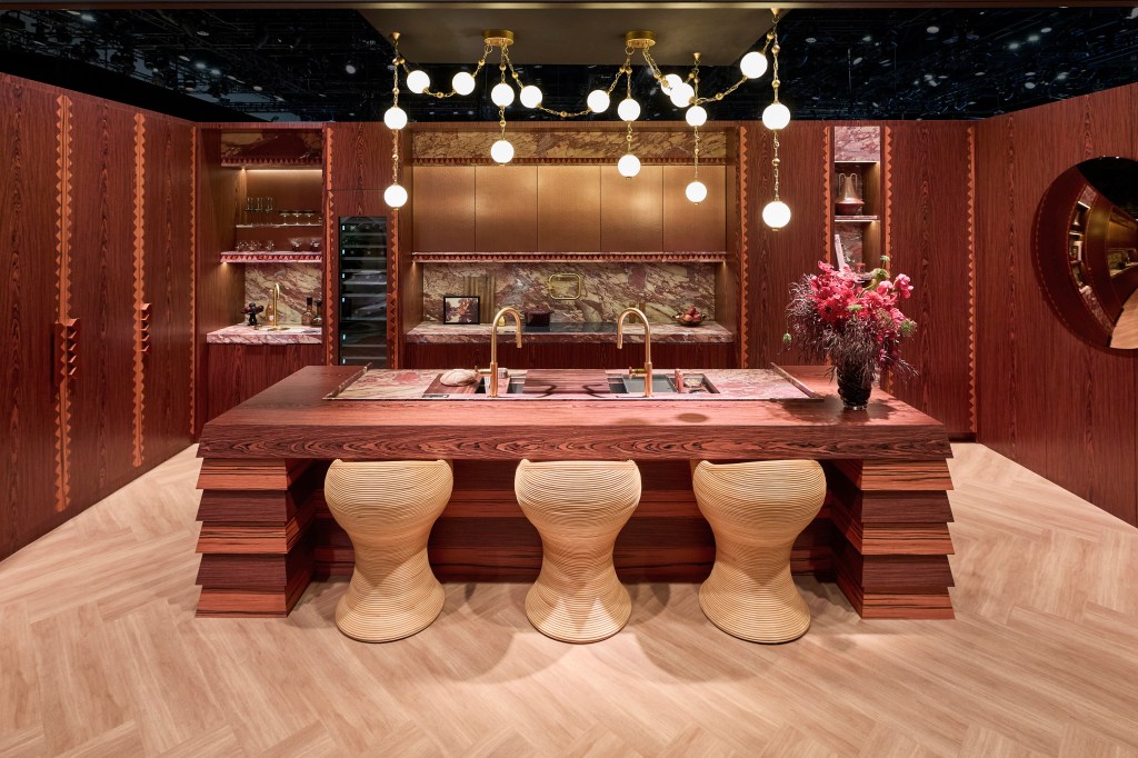 A modern kitchen featuring a large wooden island with three unique stools, a stylish sink and gold fixtures, surrounded by rich wooden cabinetry and a marble backsplash, illuminated by decorative pendant lights.