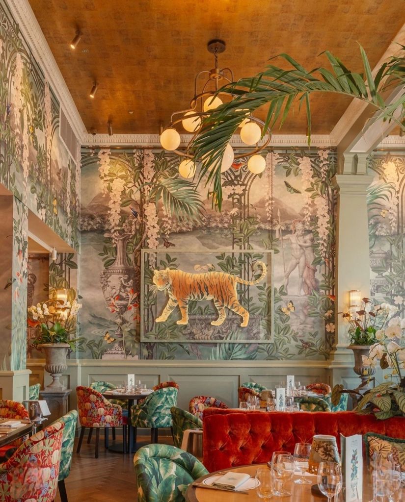 A stylish restaurant interior featuring vibrant floral-patterned chairs, a red velvet sofa, and tropical-themed wallpaper with a prominent tiger artwork. The ceiling is adorned with elegant lighting and greenery.