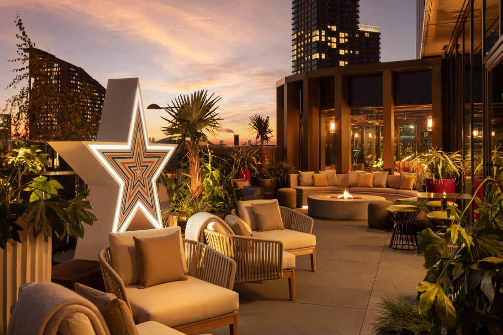 A stylish rooftop terrace featuring comfortable seating, lush greenery, and a large star-shaped light, set against a vibrant sunset with city buildings in the background.