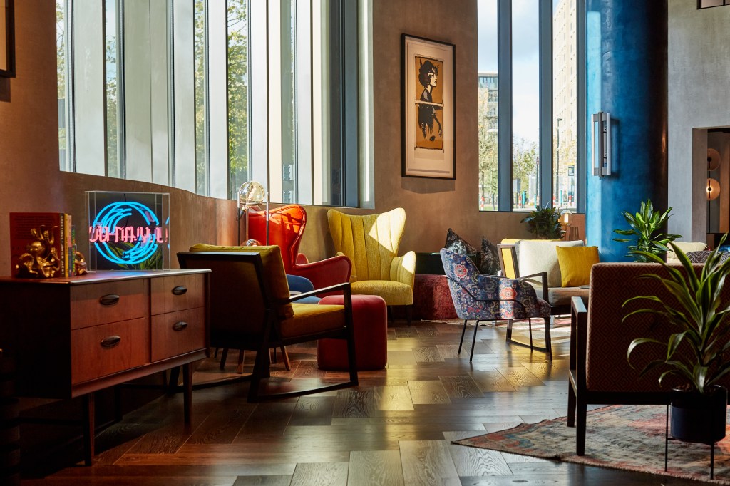 A vibrant lounge area featuring a mix of modern furniture, including colourful armchairs and a wooden sideboard. Large windows allow natural light to fill the space, highlighting decorative plants and a neon sign.