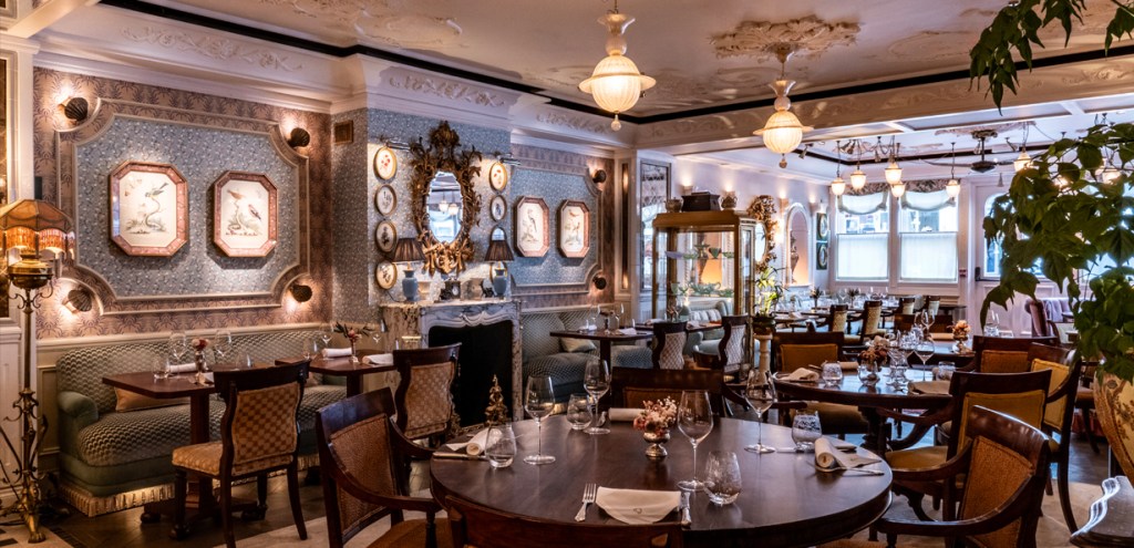Elegant restaurant interior featuring wooden tables, ornate chairs, and decorative wall art, with soft lighting and greenery.