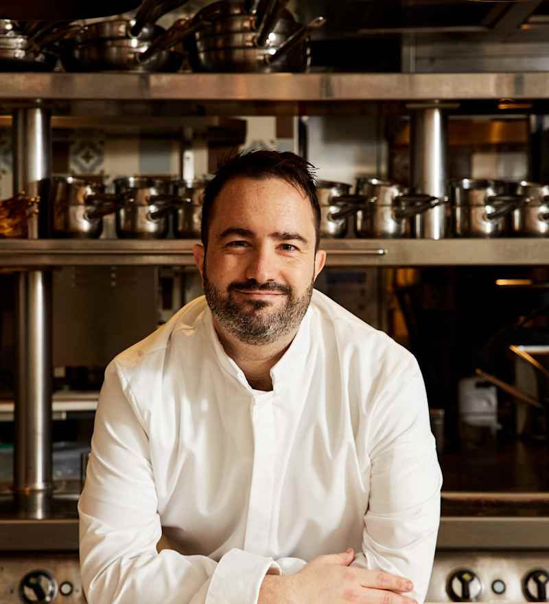 A smiling chef in a white uniform sitting in a kitchen with stainless steel cookware in the background.