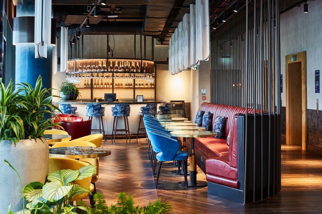 Interior of a modern bar or restaurant featuring a stylish seating area with colourful chairs, a wooden floor, and an illuminated bar in the background.