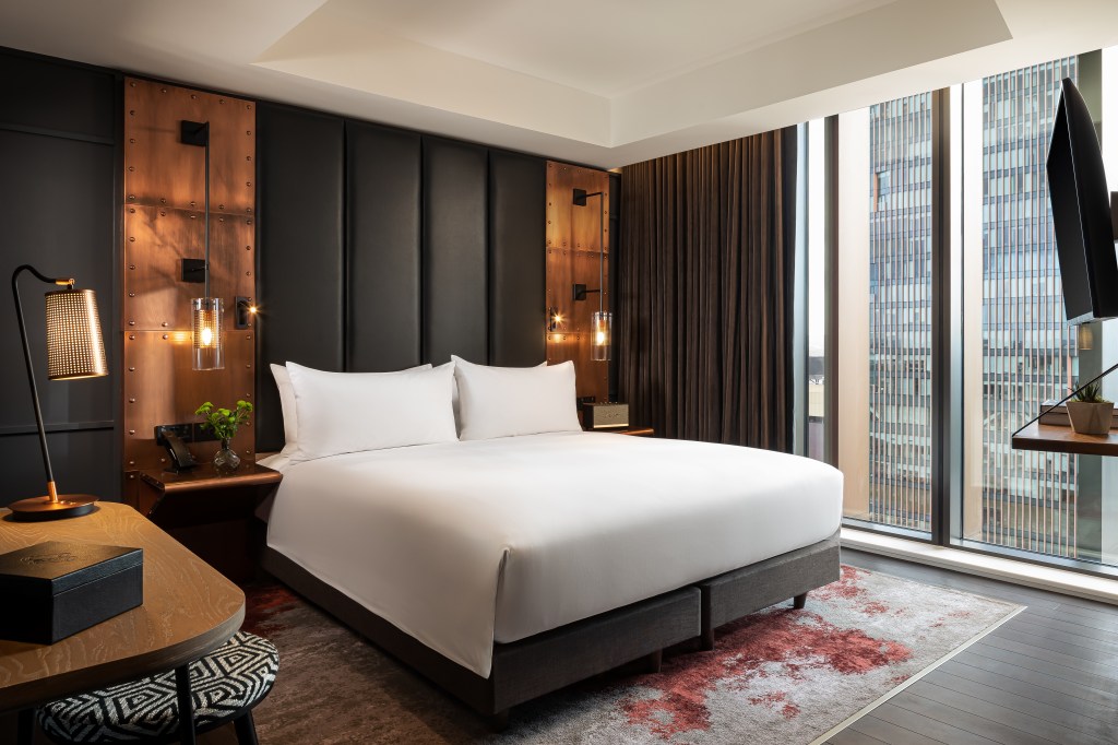 A modern hotel room featuring a large double bed with crisp white linens, stylish wooden and metal accents on the wall, and a sleek desk lamp. Large windows allow natural light to illuminate the space, which includes a minimalist decor with a patterned carpet.