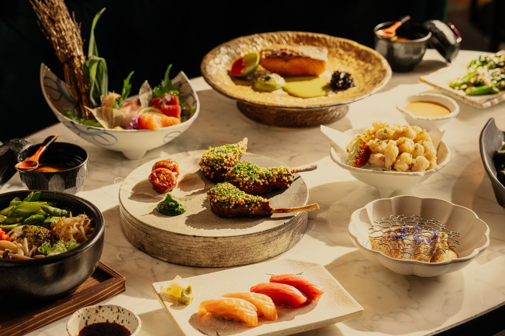 An array of beautifully presented Japanese dishes on a marble table, including sushi, sashimi, skewered items, and a bowl of vegetables.