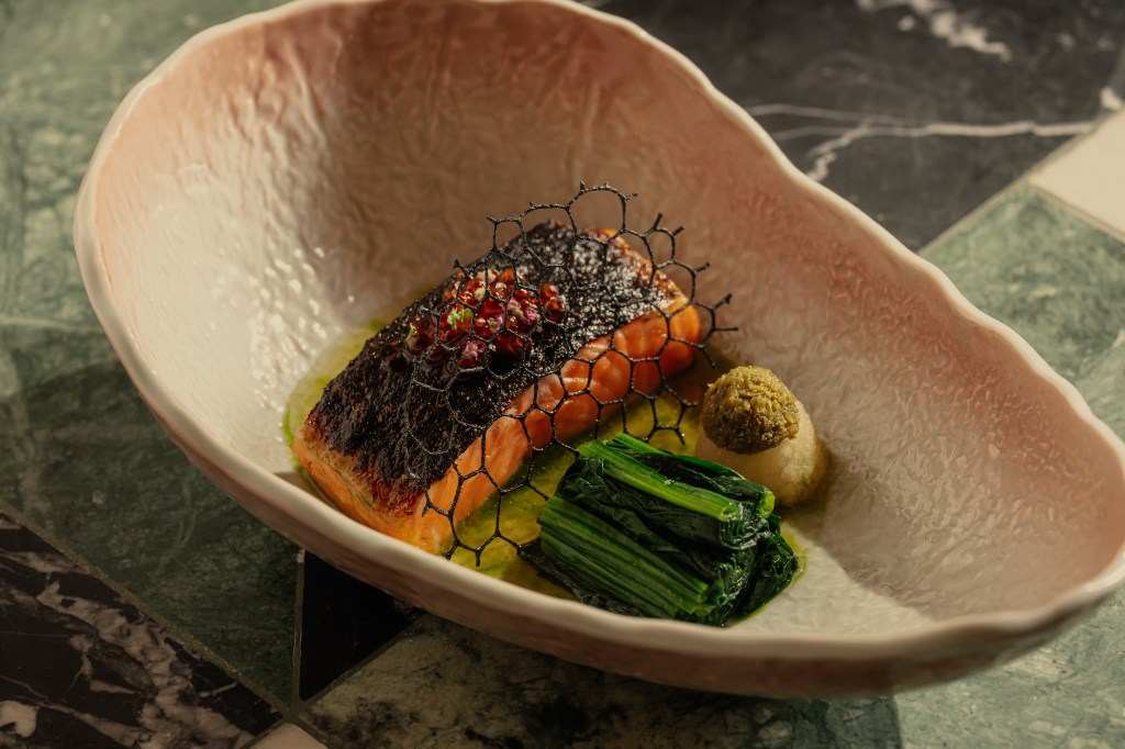 A beautifully plated dish featuring a fillet of salmon topped with a delicate crispy net, surrounded by greens and small condiments, presented in an elegant dish.