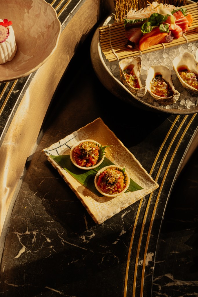 A close-up view of a decorative plate holding three small culinary dishes, each filled with a vibrant mixture, garnished with green herbs. In the background, there are oysters on a bed of ice, sushi, and a beautifully styled dessert. The setting features a dark marble surface with golden accents.