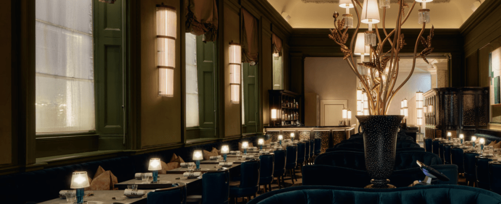 A stylish restaurant interior featuring elegant green walls, sophisticated lighting, and neatly arranged tables with blue chairs, creating a warm and inviting atmosphere.
