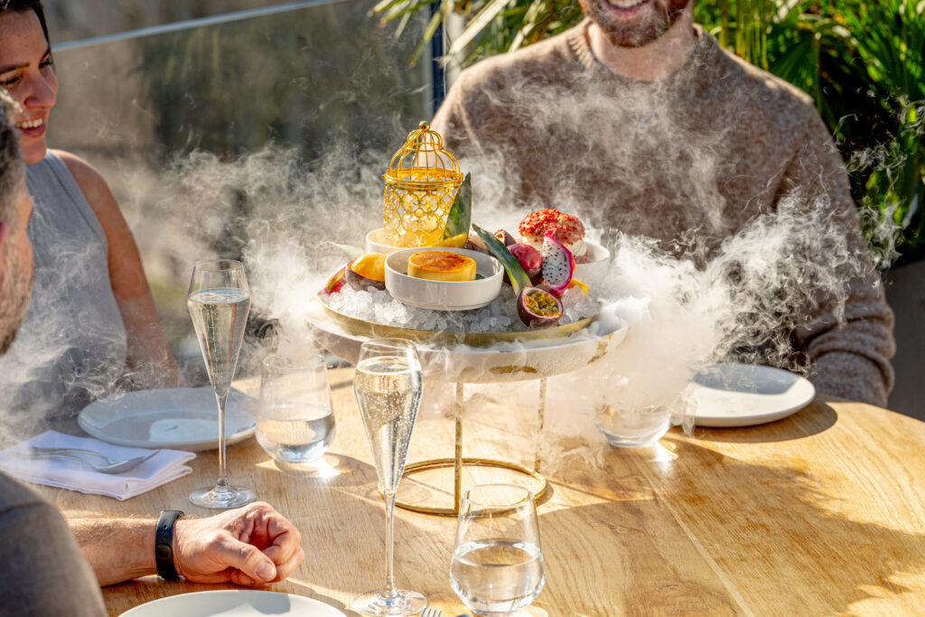 A dessert platter featuring an array of fruits including dragon fruit and passion fruit, presented on a bed of ice with steam rising around it, as friends enjoy the dessert at an outdoor dining table.