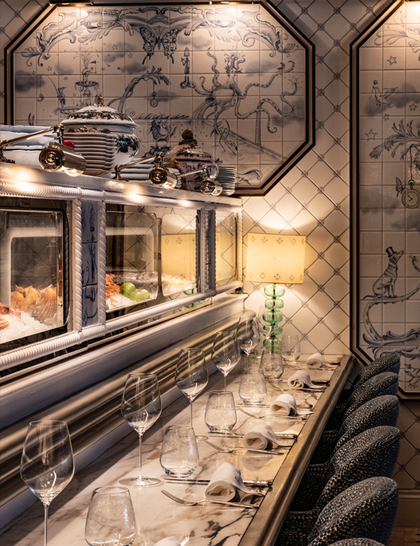 Elegant dining area featuring a marble table set with glassware and neatly folded napkins, illuminated by soft lighting. In the background, decorative tiles and a showcase displaying fresh seafood add to the sophisticated atmosphere.