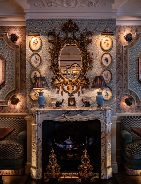 A stylish interior featuring a marble fireplace with intricate gold detailing, flanked by blue vases and decorative figurines. The walls are adorned with floral wallpaper and framed botanical prints, complemented by soft lighting.
