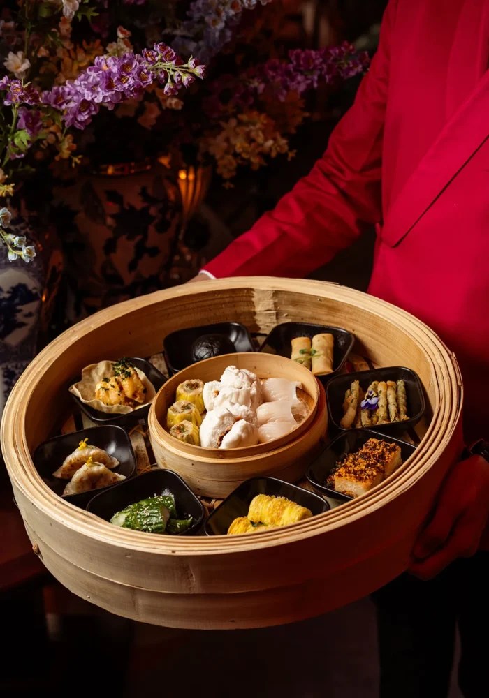 A bamboo steamer filled with a variety of dumplings and dim sum dishes, held by a person in a red shirt, with floral decorations in the background.