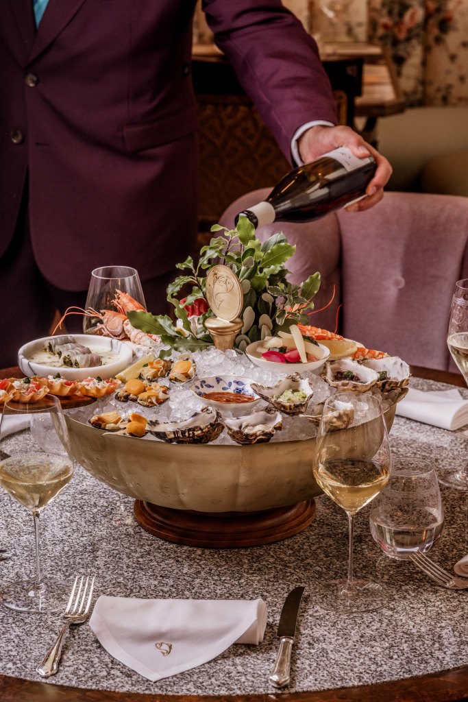A lavish seafood platter featuring oysters and shrimp, elegantly presented on ice, with a server pouring wine at a dining table adorned with glasses and silverware.