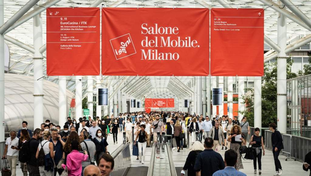 A busy exhibition hall at the Salone del Mobile in Milan, featuring numerous attendees walking under large banners displaying event information.