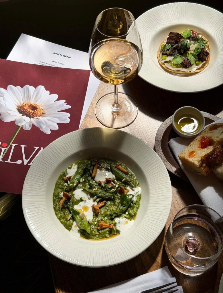A vibrant green dish featuring spinach and herbs, topped with cream and nuts, served alongside a small plate of beef tacos, a glass of white wine, and a menu with a floral design.