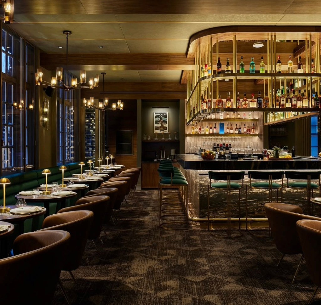 Interior of an upscale restaurant featuring elegant lighting, a stylish bar with various bottles, and wooden decor. Tables with green seating and small candle lights are arranged along the side.