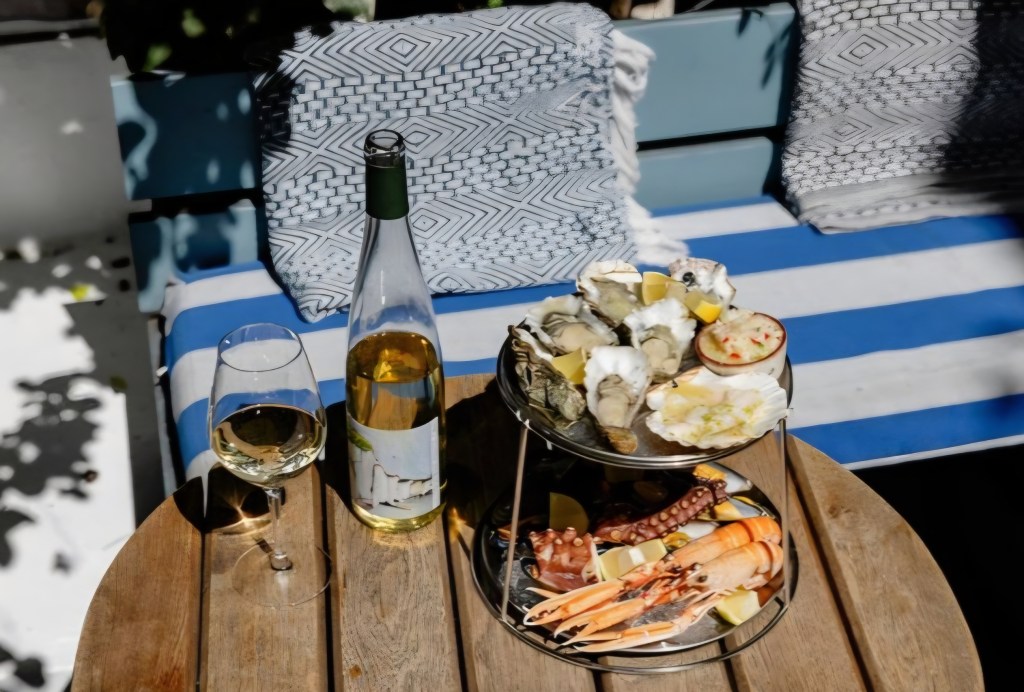 A seafood platter featuring oysters, shrimp, octopus, and scallops on a two-tier stand, accompanied by a glass of white wine and a bottle on a wooden table, with a blue and white striped background.