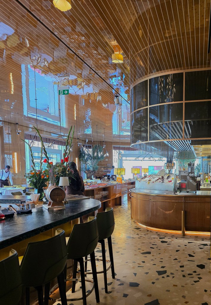 Interior of a modern restaurant featuring wooden panels, large windows, and a circular bar area with seating. Decorative plants and a well-stocked ice display are visible, along with staff attending to customers.